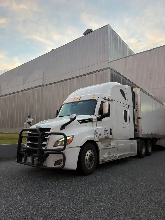 A white SDS UZ Freightliner sleeper cab with its trailer at a loading facility at dusk